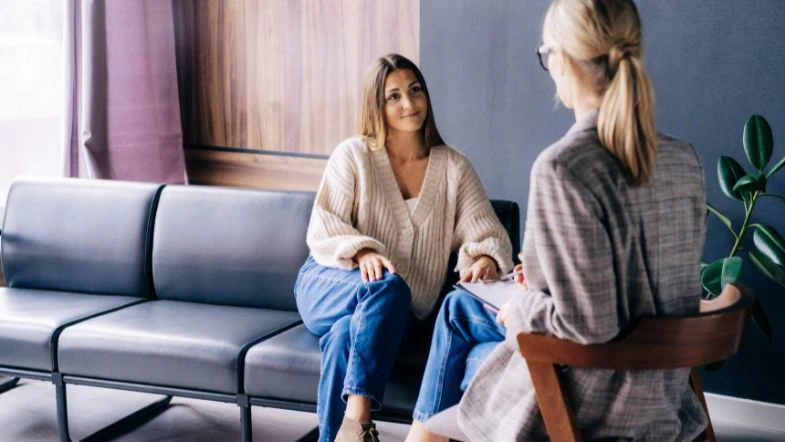 Uma mulher jovem, sentada em um sofá, conversa com uma profissional da Psicologia que está de costas. A cena ocorre em um ambiente acolhedor, sugerindo um consultório.