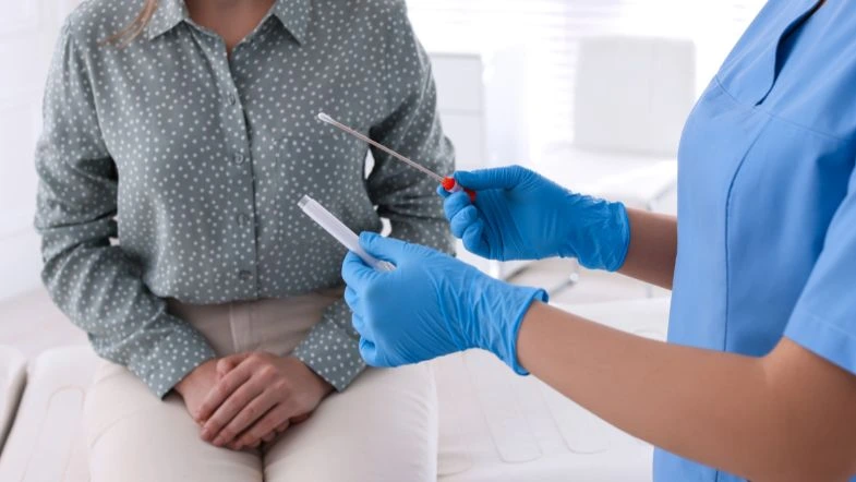 Profissional de saúde, vestindo luvas e uniforme azul, segura um swab e um tubo de coleta, preparando-se para a realização de um exame. À frente, uma paciente sentada aguarda o procedimento, demonstrando um ambiente clínico voltado para a prevenção e diagnóstico de doenças.