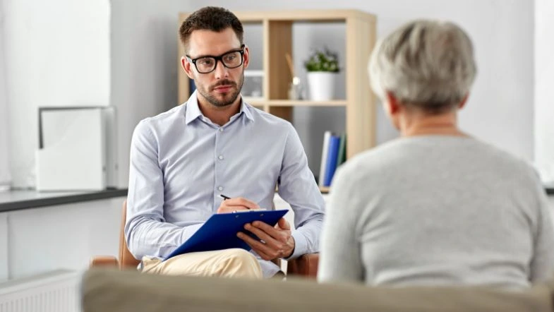 Um profissional da Psicologia está sentado em um consultório bem iluminado, segurando uma prancheta e conversando com uma paciente mais velha, que aparece de costas.