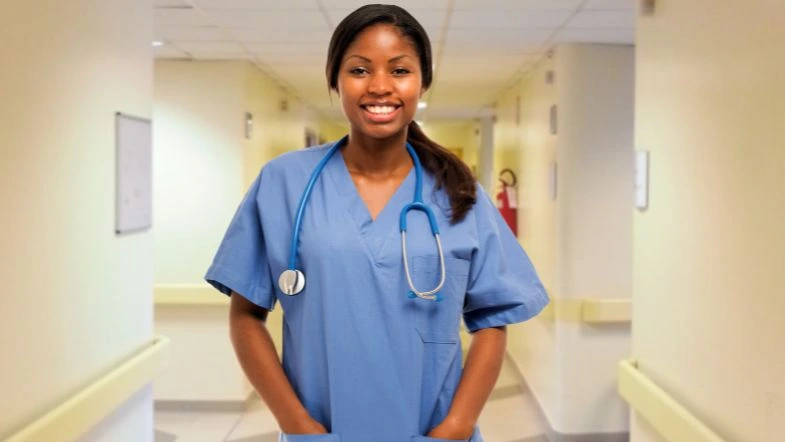 Profissional de saúde sorridente, vestindo uniforme azul e estetoscópio no pescoço, posicionada em um corredor hospitalar bem iluminado. A imagem transmite confiança, profissionalismo e acolhimento, representando a importância dos profissionais da enfermagem no cuidado com os pacientes.