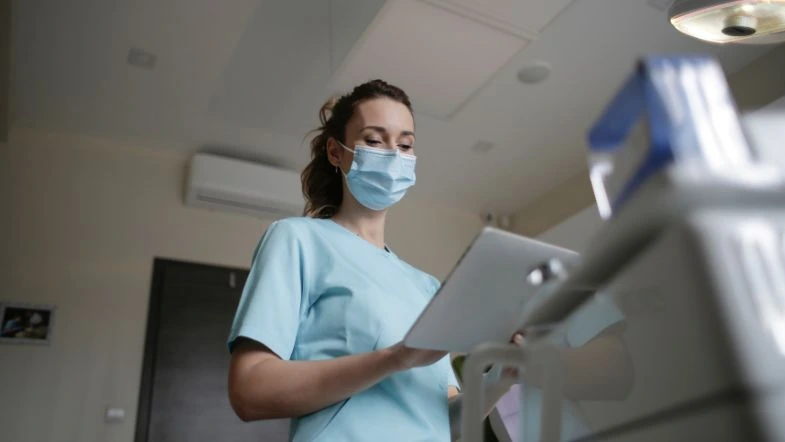 Profissional de saúde vestindo uniforme azul e máscara cirúrgica segura um tablet enquanto analisa informações médicas em um ambiente hospitalar. O cenário ao fundo sugere um ambiente clínico moderno e bem iluminado, reforçando a ideia de tecnologia e atendimento humanizado na área da saúde.