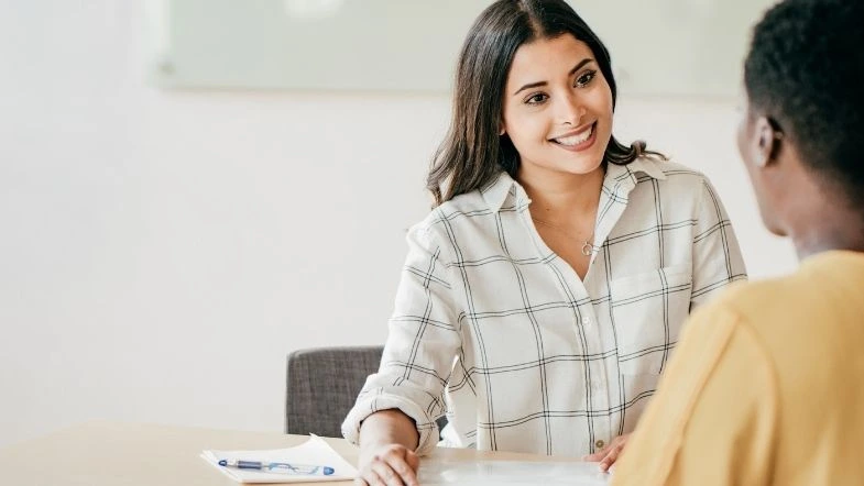 Jovem sorridente em uma conversa durante uma orientação acadêmica. A imagem representa atendimento estudantil, aconselhamento vocacional ou entrevista para ingresso no ensino superior.