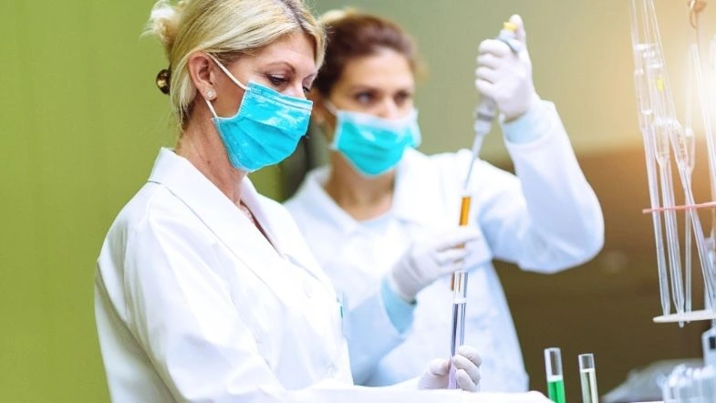 Imagem de duas cientistas em um laboratório realizando experimentos com líquidos em tubos de ensaio. Ambas usam jaleco, luvas e máscaras de proteção, garantindo a segurança nos procedimentos laboratoriais. A cena destaca a atuação feminina na ciência, especialmente na área de pesquisa e desenvolvimento em biotecnologia e saúde.