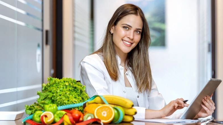 Graduação em Nutrição: o que se estuda, onde o nutricionista pode atuar e por que escolher a Faculdade ITH 6 Nutricionista sorridente com jaleco branco trabalha em um tablet enquanto está sentada à mesa com frutas, verduras e fita métrica, representando uma consulta nutricional voltada à alimentação saudável e planejamento dietético.