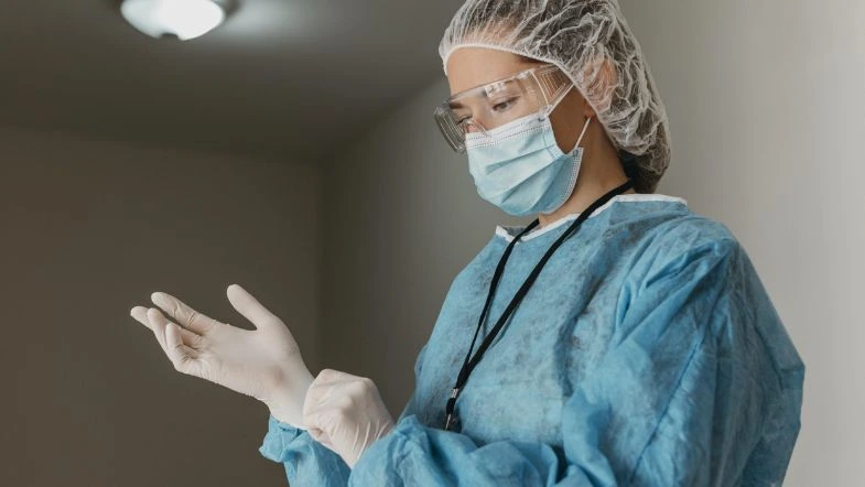 Segurança do trabalho na saúde: como prevenir acidentes 6 Profissional da saúde com máscara, touca e jaleco ajustando luvas descartáveis, demonstrando prática de segurança do trabalho.