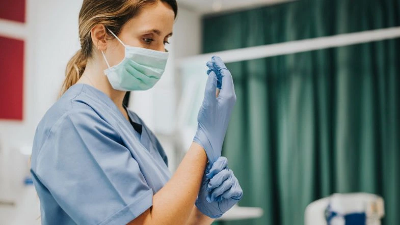 Segurança do trabalho na saúde: como prevenir acidentes 8 Profissional da saúde com máscara e luvas ajustando Equipamento de Proteção Individual em ambiente hospitalar, reforçando a segurança do trabalho.