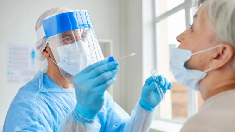 Segurança do trabalho na saúde: como prevenir acidentes 9 Profissional da saúde com EPI completo realizando teste com swab em paciente idosa, ilustrando práticas seguras no ambiente clínico.