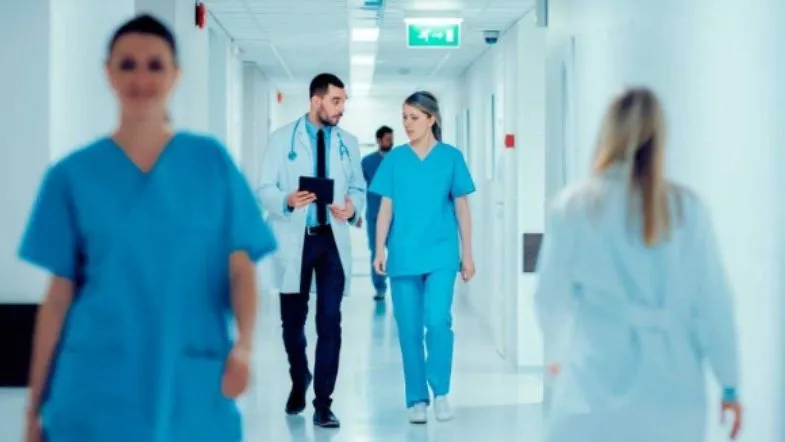 MBA em Qualidade e Acreditação em Saúde: domine ONA, JCI e ISO 6 Equipe multiprofissional da saúde caminha por corredor hospitalar, representando trabalho em equipe e ambiente clínico moderno.