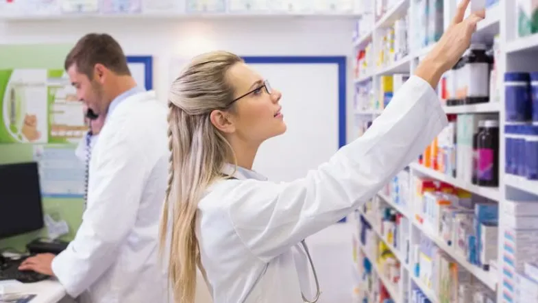 Farmácia Hospitalar: transforme a assistência clínica 6 Farmacêutica organizando medicamentos em prateleiras de uma farmácia hospitalar, enquanto um colega trabalha ao fundo.