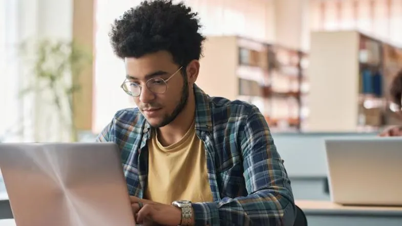 FIES 2025: como financiar sua faculdade com segurança 6 Estudante universitário com óculos e camisa xadrez estuda concentrado em um notebook dentro da biblioteca da faculdade.