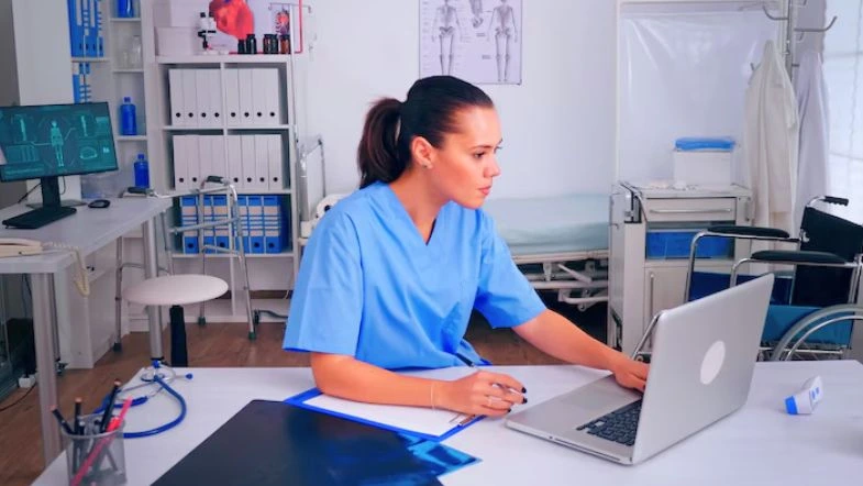 Pós-graduação em Gestão e Liderança na Saúde: destaque-se no mercado 6 Profissional da saúde vestindo jaleco azul, sentada em ambiente clínico moderno, concentrada em um notebook sobre a mesa. Ao fundo, há equipamentos médicos, cadeira de rodas e material de apoio, sugerindo um contexto de gestão hospitalar ou análise clínica.