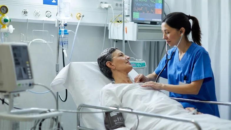 UTI na prática: tudo sobre salário e perfil do enfermeiro intensivista 6 Enfermeira sorridente auscultando paciente idosa internada em leito de UTI, demonstrando cuidado humanizado e monitoramento contínuo.