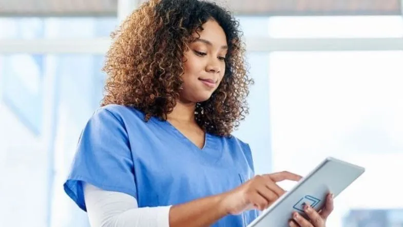 Compliance em Saúde: o que é e quanto ganha esse profissional 6 Profissional da enfermagem com jaleco azul utilizando tablet digital em ambiente hospitalar, representando tecnologia aplicada à gestão.