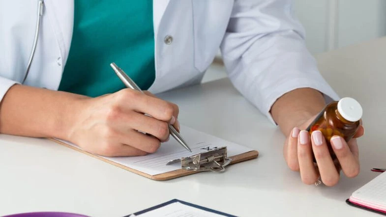 Farmácia: o que faz um farmacêutico, onde atuar e como se destacar na área da saúde 6 Profissional da saúde com jaleco branco segura frasco de medicamento enquanto escreve em uma prancheta, representando prescrição farmacêutica e atenção clínica.