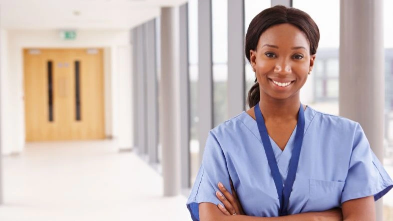 Gestão comercial na saúde: como impulsionar a inovação 6 Enfermeira sorridente de braços cruzados em corredor iluminado de hospital, vestindo uniforme azul-claro, representando confiança, liderança e profissionalismo na área da saúde.