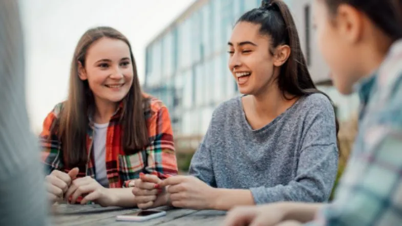 Enem: o que é, como funciona, quem pode fazer e como usar no Sisu, Prouni e outros programas 7 Três adolescentes conversam e sorriem ao ar livre, demonstrando integração social e ambiente escolar descontraído.