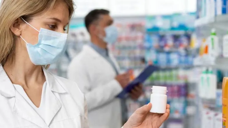 Farmácia Hospitalar: transforme a assistência clínica 7 Farmacêutica com máscara facial avaliando um frasco de medicamento, com farmacêutico ao fundo conferindo anotações.