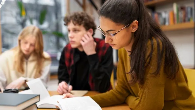 FIES 2025: como financiar sua faculdade com segurança 7 Aluna com óculos estuda atentamente um livro em uma mesa de estudos, com dois colegas ao fundo revisando conteúdos.