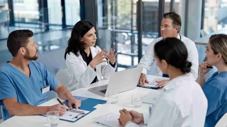 MBA em Gestão Hospitalar: transforme sua carreira na administração da saúde 7 Profissionais da saúde em reunião ao redor de uma mesa, com uma mulher de jaleco liderando a discussão. O grupo está em um ambiente corporativo e moderno, com anotações, pastas e um notebook, simbolizando gestão e planejamento hospitalar.
