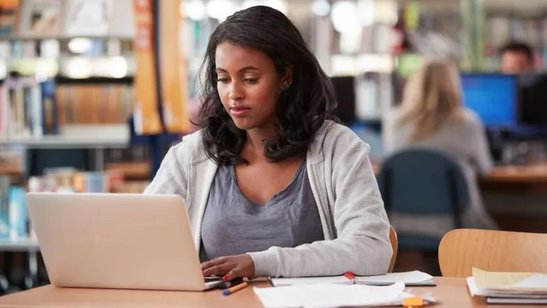 Graduação: por que ela é essencial para sua carreira? 7 Aluna concentrada estuda em biblioteca universitária utilizando notebook e cadernos, ilustrando o ambiente acadêmico e a rotina de quem está cursando uma graduação.