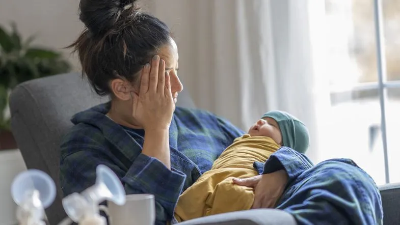 Cuidados no pós-parto: aula gratuita sobre assistência à saúde da mulher puérpera 7 Mãe com expressão de exaustão segura o bebê recém-nascido nos braços, retratando os desafios emocionais do pós-parto.