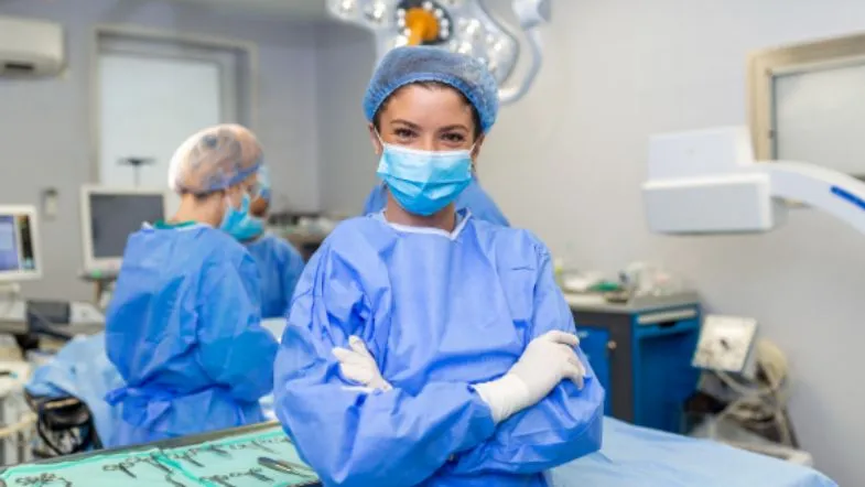 MBA em Qualidade e Acreditação em Saúde: domine ONA, JCI e ISO 8 Enfermeira sorri com braços cruzados em centro cirúrgico, usando roupa estéril e máscara, enquanto outros profissionais operam ao fundo.