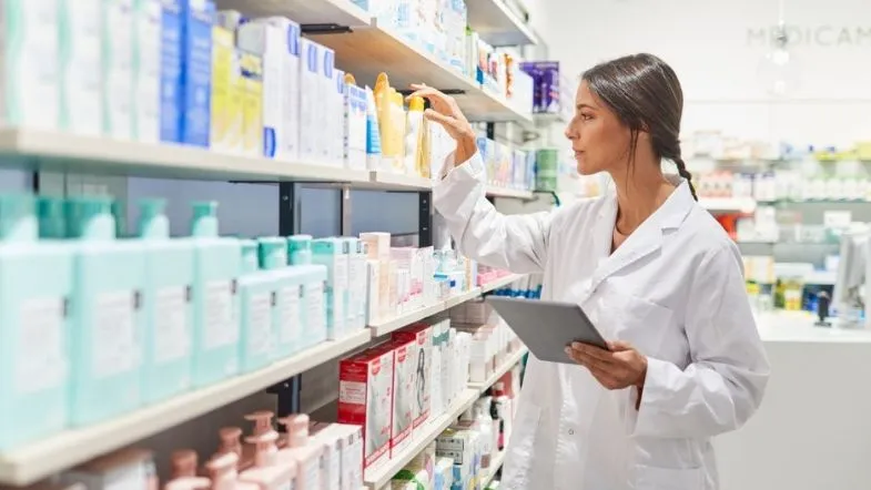 Farmácia Hospitalar: transforme a assistência clínica 8 Profissional da farmácia inspecionando produtos em uma prateleira, segurando um tablet para controle de estoque.