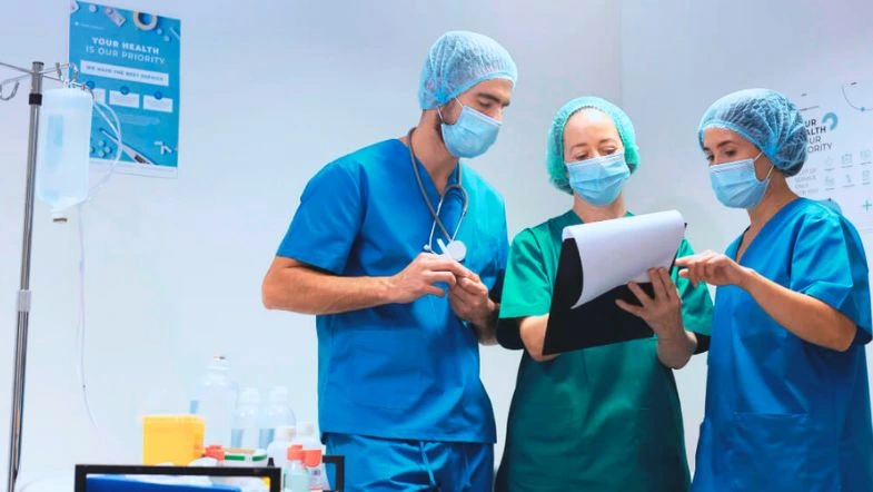 Pós-graduação em Gestão e Liderança na Saúde: destaque-se no mercado 8 Três profissionais da saúde em ambiente hospitalar, usando roupas cirúrgicas e máscaras, revisam informações em uma prancheta ao lado de equipamentos médicos e suporte de soro. A cena representa trabalho em equipe, segurança e atenção aos protocolos clínicos.