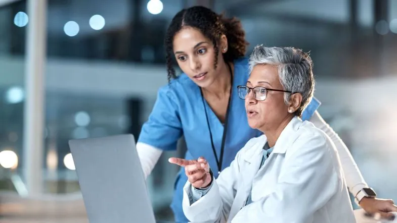 MBA em Gestão Hospitalar: transforme sua carreira na administração da saúde 8 Duas profissionais da saúde analisam informações em um laptop em ambiente hospitalar. Uma mulher mais jovem, com uniforme azul, observa atentamente enquanto a outra, mais experiente, com cabelo grisalho e jaleco branco, aponta para a tela durante uma conversa técnica.