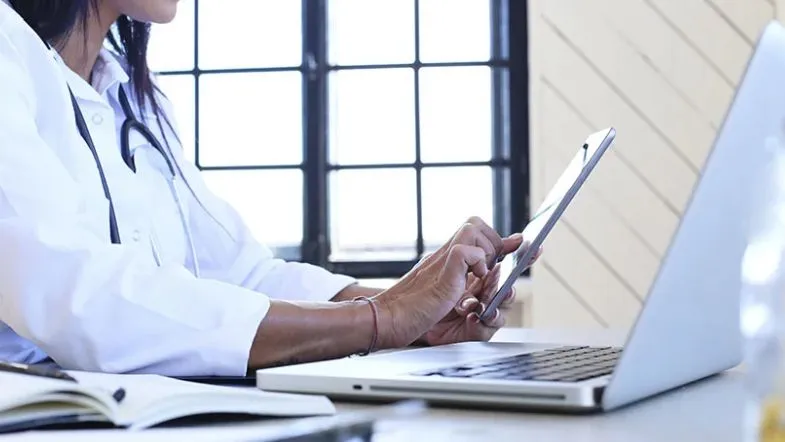 Marketing digital para profissionais da saúde: participe do evento gratuito que vai transformar sua presença online 7 Profissional da saúde usando tablet ao lado de um notebook em ambiente iluminado, representando a digitalização e o uso de tecnologia na rotina clínica.