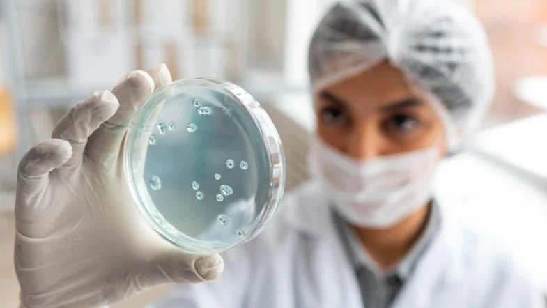 Biomedicina: avanços tecnológicos na saúde 9 Profissional de laboratório com touca, máscara e luvas observando uma placa de Petri com culturas bacterianas, representando análises microbiológicas na biomedicina.