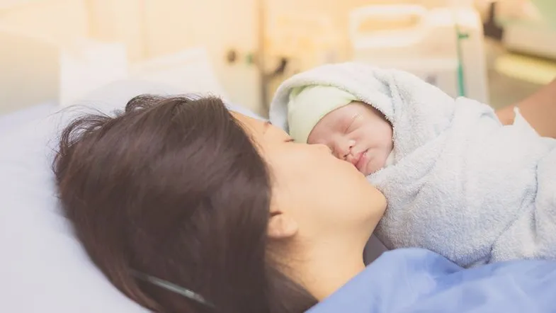 Cuidados no pós-parto: aula gratuita sobre assistência à saúde da mulher puérpera 8 Mãe beija o bebê enrolado em manta logo após o parto, ainda no ambiente hospitalar, representando vínculo pós-parto.