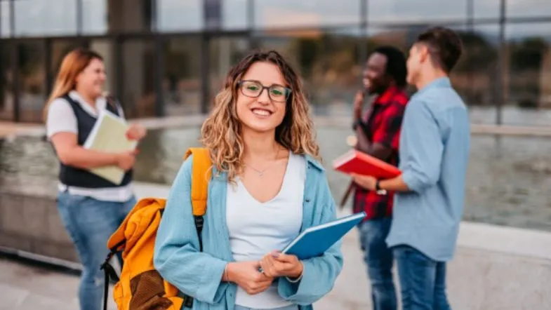 Enem: o que é, como funciona, quem pode fazer e como usar no Sisu, Prouni e outros programas 9 Estudante universitária segura cadernos e sorri, com colegas conversando ao fundo em campus universitário moderno.