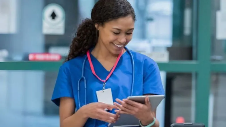 MBA em Qualidade e Acreditação em Saúde: domine ONA, JCI e ISO 9 Enfermeira com crachá e estetoscópio sorri enquanto utiliza tablet, representando profissionais especializados pelo MBA em Qualidade e Acreditação.