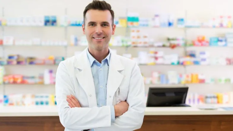 Farmácia Hospitalar: transforme a assistência clínica 9 Farmacêutico sorridente de braços cruzados em frente ao balcão de uma farmácia, com prateleiras de medicamentos ao fundo.