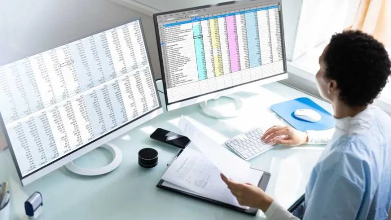 Auditoria em Serviços de Saúde: alavanque sua carreira 9 Auditor da área da saúde analisando planilhas com dados financeiros e operacionais em duas telas de computador.