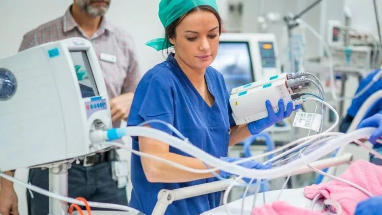 UTI na prática: tudo sobre salário e perfil do enfermeiro intensivista 9 Técnica de enfermagem ajustando bomba de infusão em leito de UTI neonatal, cercada por monitores e equipamentos de suporte ao paciente crítico.