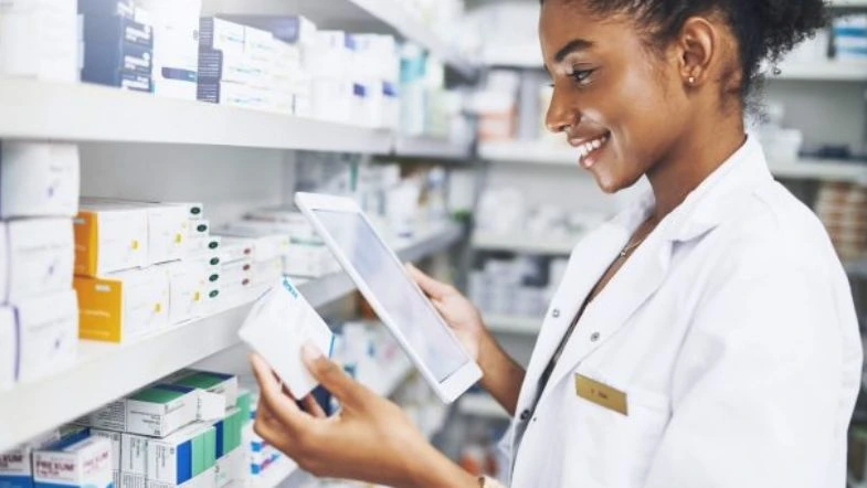 Farmácia: o que faz um farmacêutico, onde atuar e como se destacar na área da saúde 9 Farmacêutica sorridente analisa uma embalagem de medicamento enquanto consulta informações em um tablet, em meio a prateleiras repletas de caixas de remédios em uma farmácia.