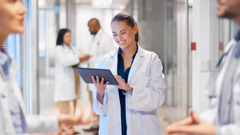 MBA em Gestão Hospitalar: transforme sua carreira na administração da saúde 9 Profissional da saúde sorridente utilizando um tablet em um corredor de hospital, vestida com jaleco branco. Ao fundo, outros profissionais também interagem em ambiente clínico moderno, sugerindo trabalho colaborativo e gestão hospitalar eficiente.