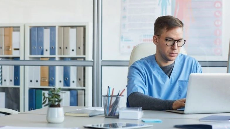 Gestão comercial na saúde: como impulsionar a inovação 9 Profissional da saúde com jaleco azul e óculos trabalha em frente ao notebook, em ambiente administrativo com pastas e materiais de escritório ao fundo.