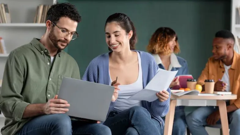 Ensino superior em 2025: saiba como ingressar na faculdade com segurança 6 Dois estudantes analisam documentos e utilizam notebook em sala de aula, simbolizando o planejamento para entrada na faculdade.