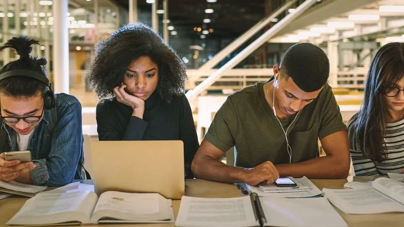 Graduação aumenta em até 57% o salário no brasil: veja como começar a sua 6 Jovens universitários estudando juntos em biblioteca, usando laptop e cadernos, representando dedicação à graduação.