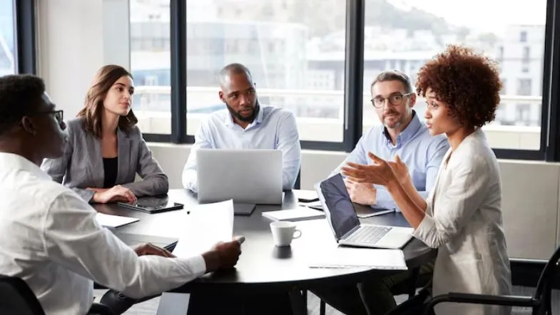 Graduação em Recursos Humanos: guia completo para impulsionar resultados 6 Equipe de profissionais reunidos em sala de reunião moderna, discutindo estratégias com laptops e documentos sobre a mesa.