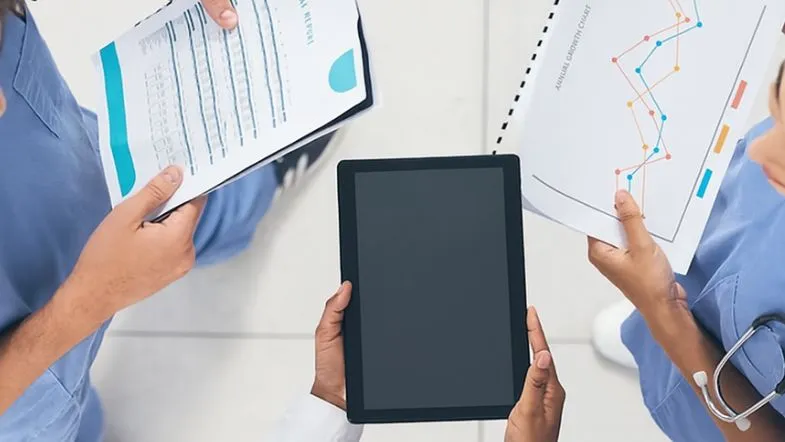 Três profissionais da saúde vistos de cima, analisando relatórios gráficos e financeiros enquanto seguram uma prancheta, papéis e um tablet, simbolizando trabalho colaborativo e gestão de dados em ambiente hospitalar.