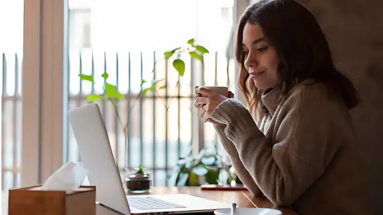 Curso híbrido ou EAD? Veja as diferenças e qual é melhor para você 6 Estudante em ambiente confortável assiste a conteúdo digital com uma xícara na mão, destacando o equilíbrio entre rotina e aprendizado no ensino híbrido.
