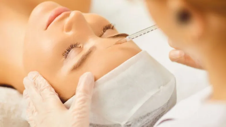 Estética em 3 dimensões: participe da aula gratuita e eleve seus resultados 6 Profissional da saúde estética realizando aplicação de procedimento injetável na região da testa de uma paciente deitada, com touca descartável e luvas de proteção.