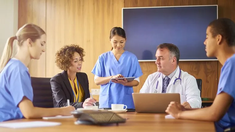 Reunião entre profissionais de saúde e gestores hospitalares em uma sala corporativa, com laptops e cadernos à mesa, ilustrando a tomada de decisões em equipe multidisciplinar.