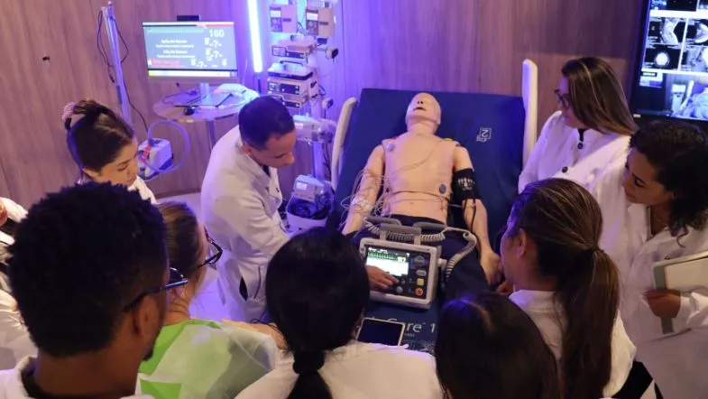Urgência e Emergência na prática: alunos da pós ITH vivem imersão no centro Hospcom 7 Grupo de alunos da pós ITH interagindo com manequim de alta fidelidade durante simulação de parada cardíaca em ambiente tecnológico do centro Hospcom.