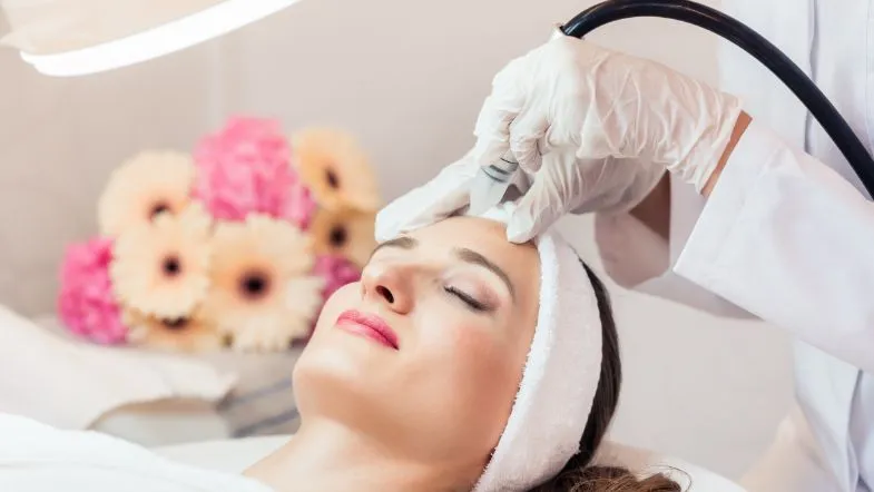 Estética em 3 dimensões: participe da aula gratuita e eleve seus resultados 7 Paciente deitada em ambiente clínico, recebendo tratamento estético facial com aparelho, ao fundo flores decorativas e iluminação adequada.