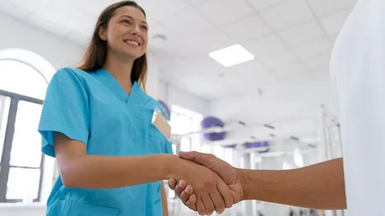 Profissional da saúde sorridente com uniforme azul cumprimentando alguém com um aperto de mão, representando boas práticas de relacionamento e acolhimento na gestão hospitalar.